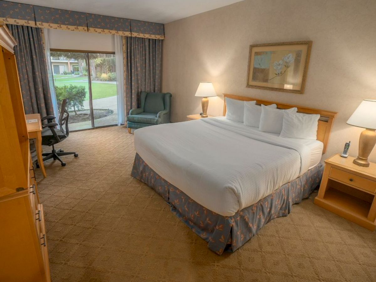 A hotel room features a neatly made bed, two nightstands with lamps, a desk with a chair, and a sliding door leading to a patio area.