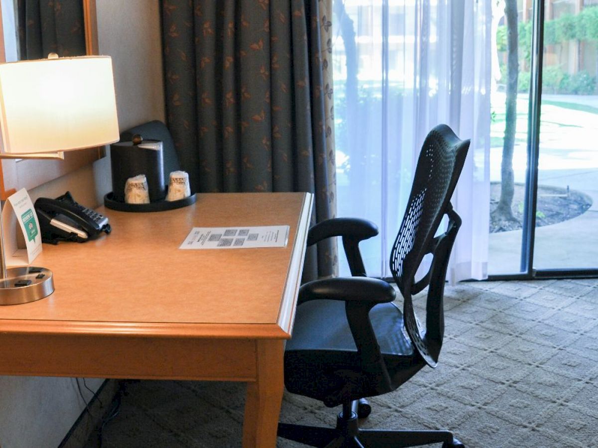 A desk with a lamp, phone, coffee station, and an office chair is set in front of a window overlooking a patio area with curtains.