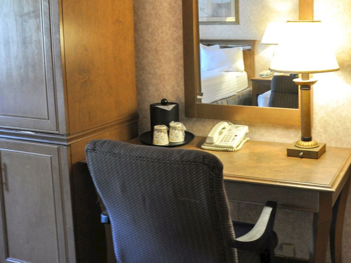 A hotel room setup with a desk, chair, lamp, telephone, cups, and a wardrobe, along with a mirror reflecting part of a bed with pillows.