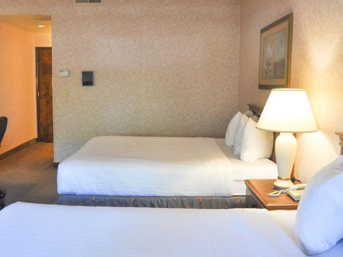 The image shows a hotel room with two beds, a side table with a lamp, a picture frame on the wall, and a desk with a chair.