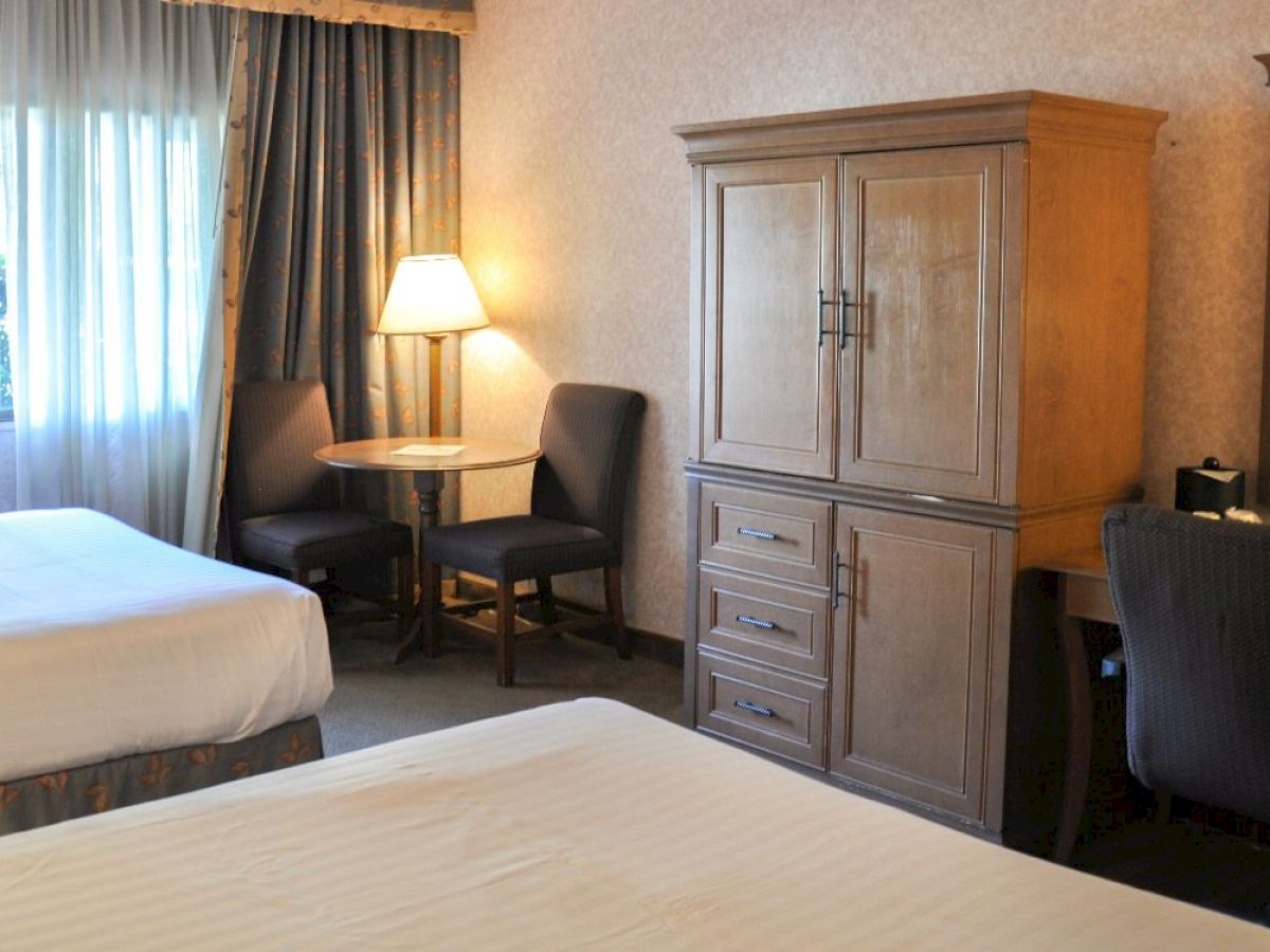 The image shows a hotel room with two beds, a wooden armoire, a round table with two chairs, a lamp, and a desk with a mirror and chair.