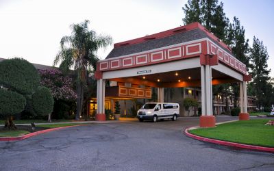 A white van is parked under the entrance canopy of a building with well-maintained landscaping and trees surrounding it.