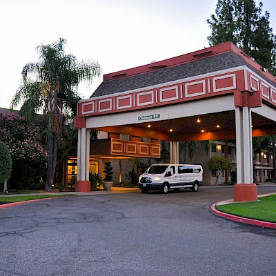 A white van is parked under the entrance canopy of a building with well-maintained landscaping and trees surrounding it.