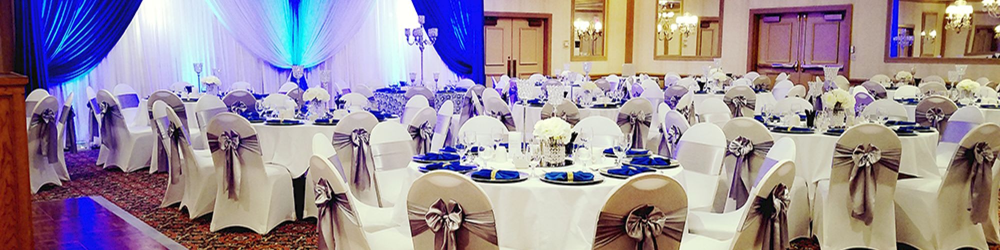 A beautifully decorated banquet hall with round tables, white chair covers, silver ribbons, chandeliers, and blue draped backdrop.