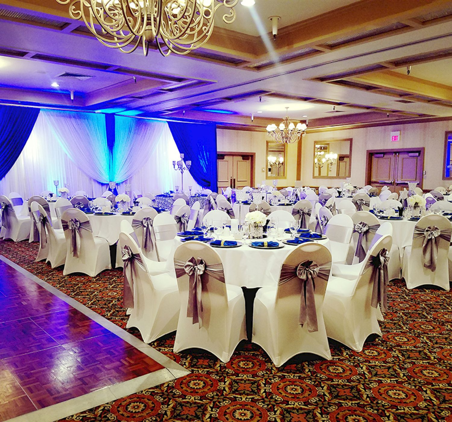 The image shows an elegantly decorated banquet hall with round tables covered in white cloth, adorned with chair bows and blue drapes, ready for an event.