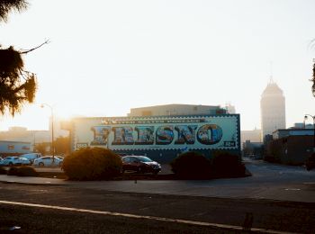 The image shows a parking lot and a large sign with the word "Fresno," with buildings and a tall tower in the background during a sunrise or sunset.