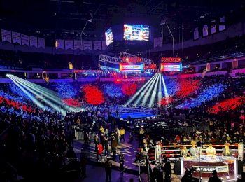 A large arena is filled with spectators for a boxing event. Spotlights illuminate the ring while the crowd is bathed in red and blue lights.