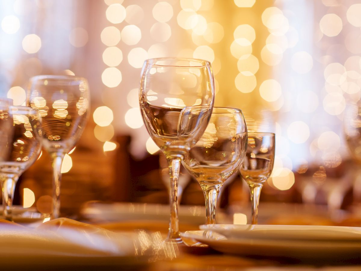 The image shows an elegant table setting with glasses and plates, with a blurred background featuring warm, bokeh lights for a festive atmosphere.