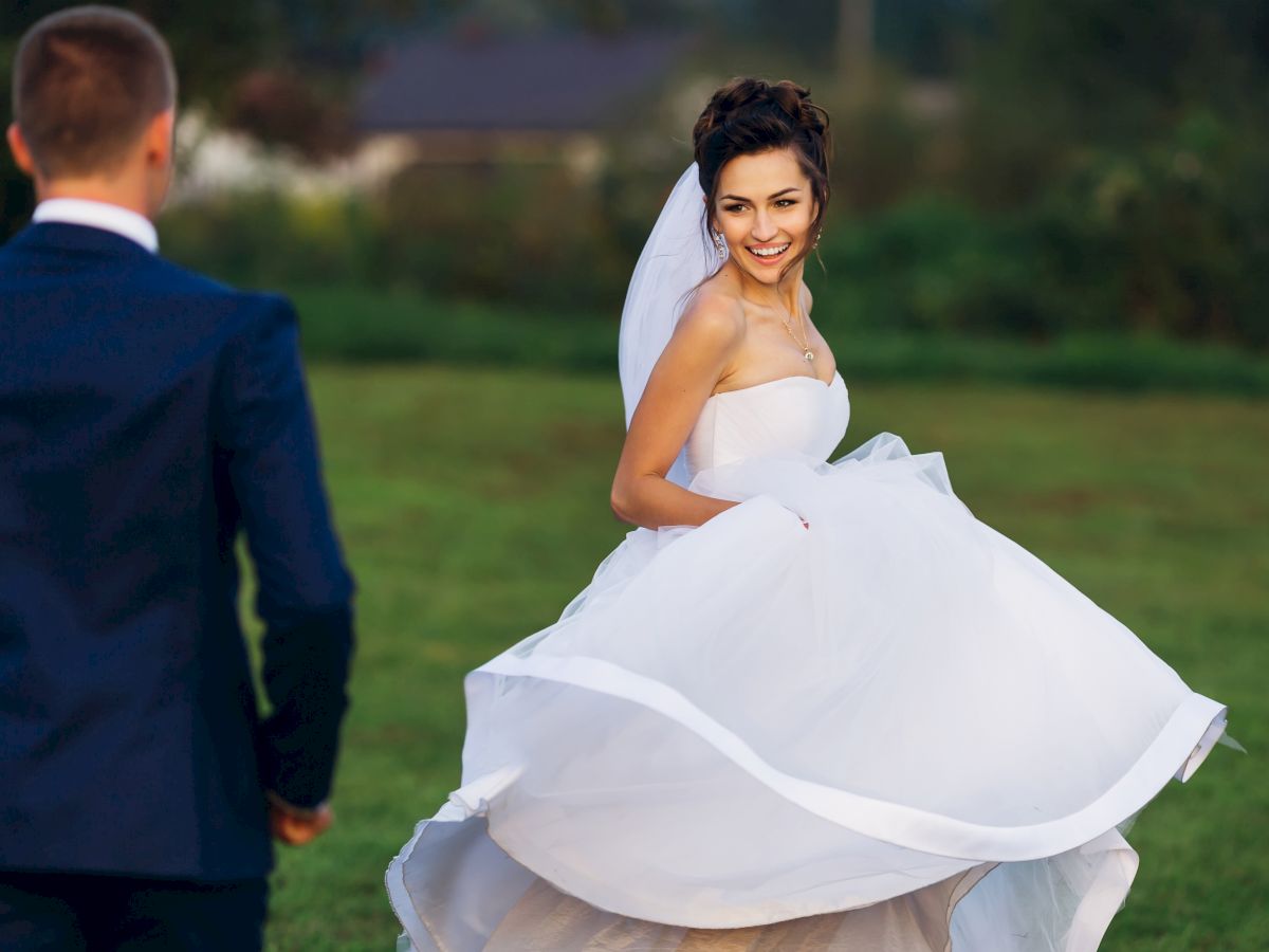 A bride in a white wedding dress twirls on a grassy field while smiling at a man in a dark suit approaching her.
