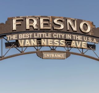 The image shows an archway sign that reads "Fresno, the best little city in the U.S.A., Van Ness Ave., Entrance," against a clear sky.