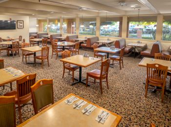 The image shows an empty restaurant dining area with neatly set wooden tables and chairs, and windows overlooking a parking lot.