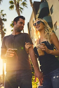 A man and a woman are walking outside with drinks in hand, smiling at each other, surrounded by palm trees and a building.