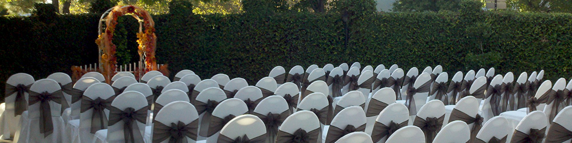 An outdoor event setup with rows of chairs adorned with white covers and dark bows, facing an arch decorated with flowers, surrounded by greenery.