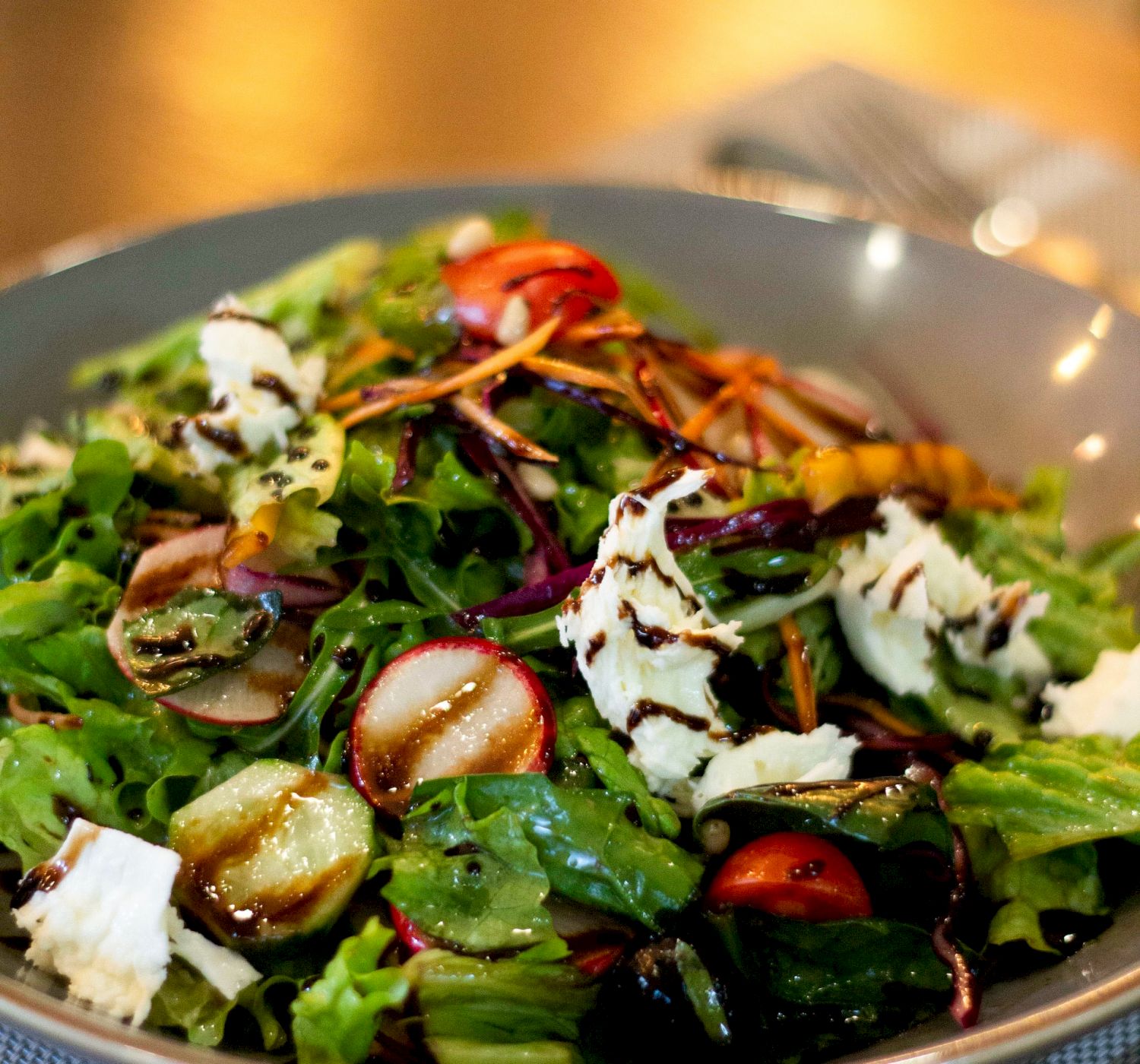 A mixed green salad with cherry tomatoes, sliced vegetables, and cheese, drizzled with a balsamic glaze, served in a gray bowl.