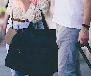 Two people are walking down a street with luggage, suggesting they are traveling or returning from a trip, carrying a duffel bag and a suitcase.