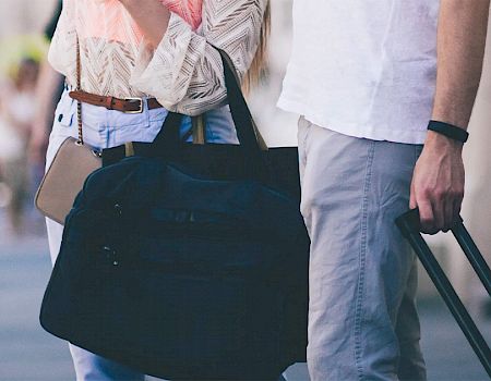 Two people are walking down a street with luggage, suggesting they are traveling or returning from a trip, carrying a duffel bag and a suitcase.