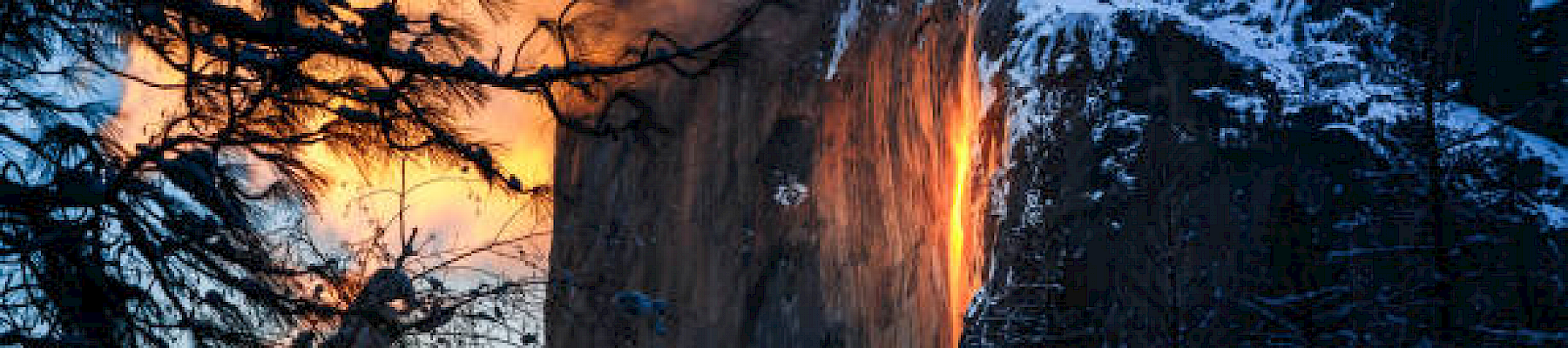 This image shows a sunset illuminating a waterfall on a cliff, making it appear as if it's flowing with fire, surrounded by snow-covered trees and mountains.