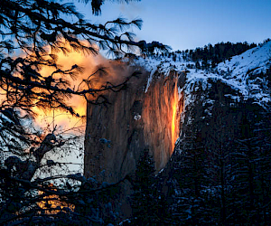 This image shows a sunset illuminating a waterfall on a cliff, making it appear as if it's flowing with fire, surrounded by snow-covered trees and mountains.