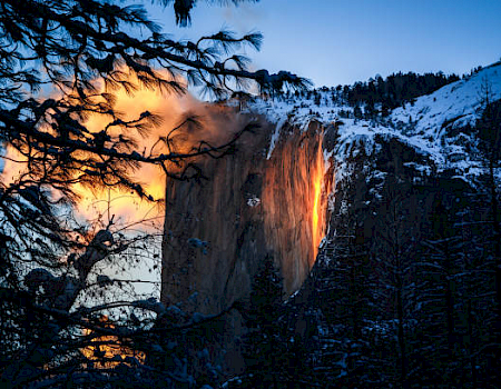 This image shows a sunset illuminating a waterfall on a cliff, making it appear as if it's flowing with fire, surrounded by snow-covered trees and mountains.