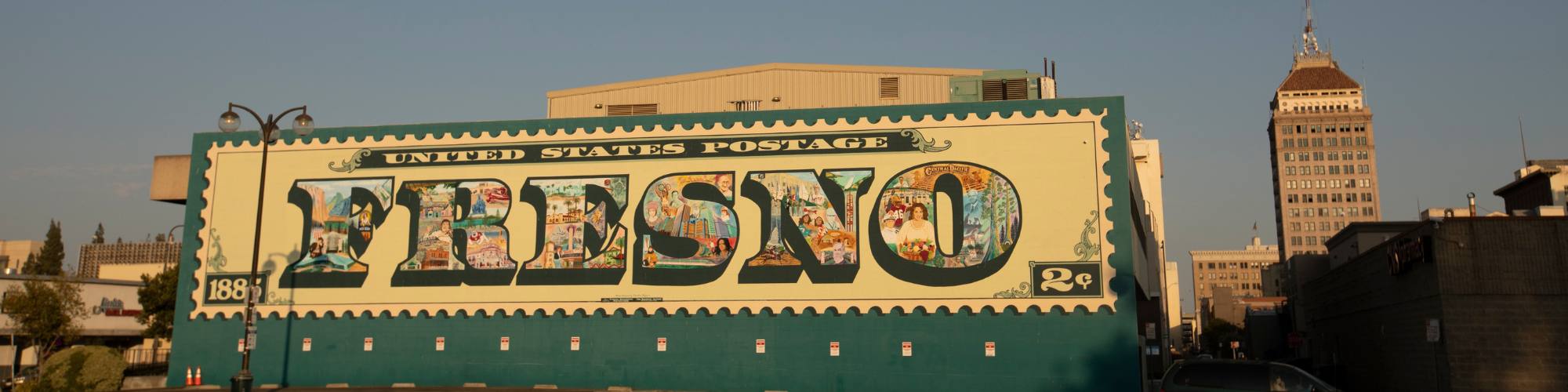 A mural depicting a large postage stamp with the text "Fresno" is painted on a building wall in an urban area with another building in the background.