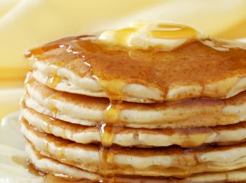 A stack of pancakes topped with butter and drizzled with syrup on a white plate, set against a soft yellow background.