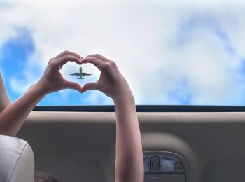 A pair of hands forms a heart shape against a background of a plane flying in the sky, visible through a car's sunroof.