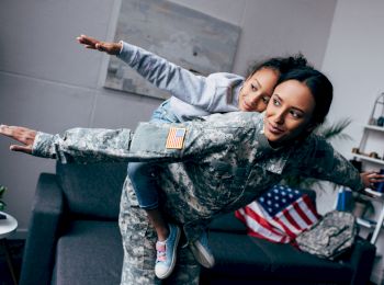 A woman in military camouflage playfully carries a child on her back, both with arms outstretched, in a room with a flag and couch.