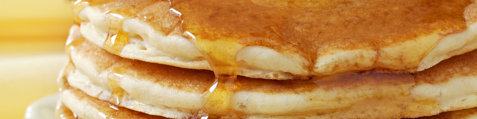 The image shows a stack of pancakes topped with melting butter and drizzled with syrup, arranged attractively on a plate.