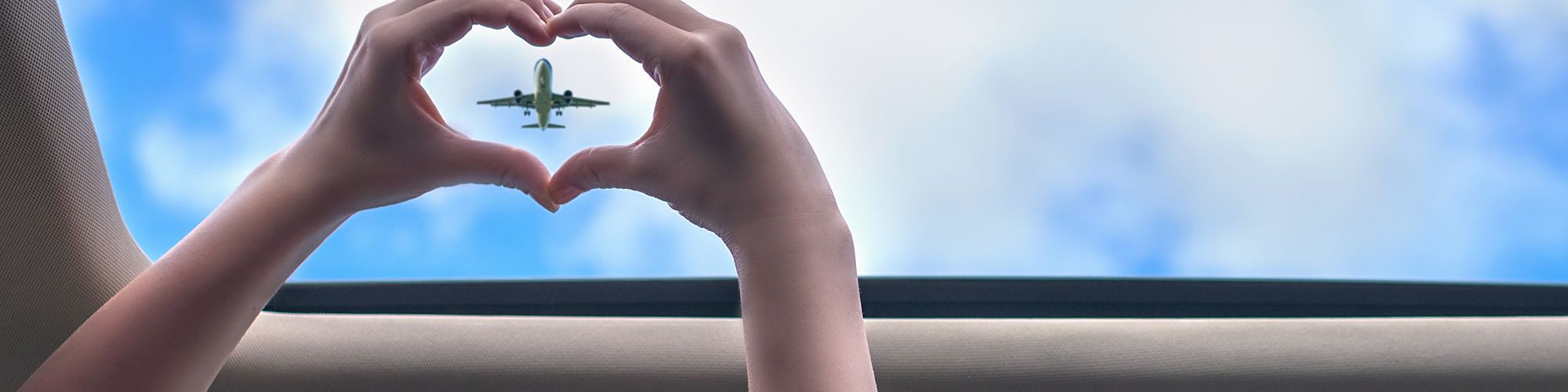 A person forms a heart shape with their hands, framing an airplane in the sky, viewed through the sunroof of a car.