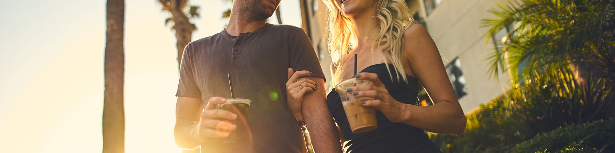 A man and woman walking outside, smiling at each other, holding drinks, with palm trees and sunlight in the background.