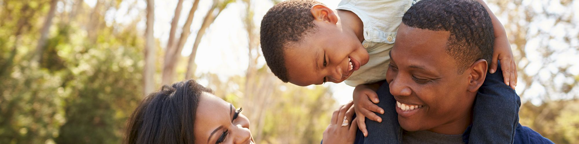 A happy family is enjoying time outdoors; a man holds a child on his shoulders while a woman stands beside them, all smiling.