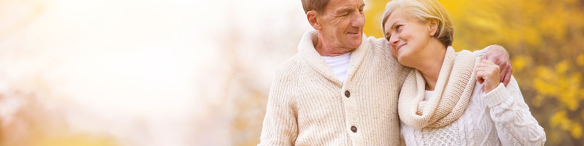 A senior couple, dressed in cozy sweaters, walking arm-in-arm through a picturesque autumn landscape with golden foliage and a soft, glowing light.