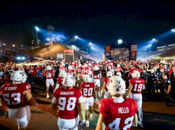 Football players in red and white uniforms walk towards a crowded stadium under night lights, preparing for a game.