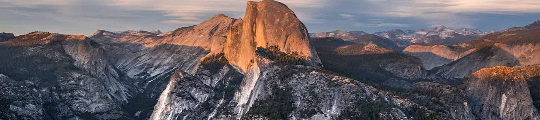 The image shows a stunning view of Half Dome, a prominent rock formation in Yosemite National Park, with surrounding mountains and a partly cloudy sky.