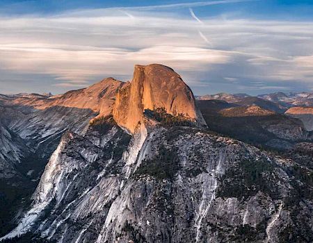 The image shows a stunning view of Half Dome, a prominent rock formation in Yosemite National Park, with surrounding mountains and a partly cloudy sky.