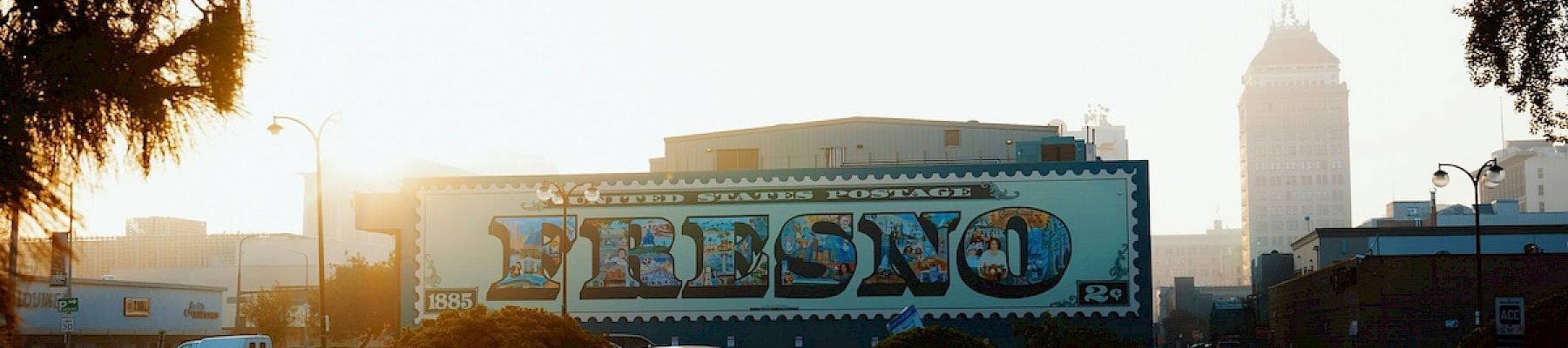 A city street scene during sunrise with a large mural reading "Fresno," cars parked beside it, and a tall building in the background, ending the sentence.