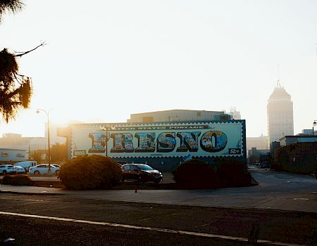 A city street scene during sunrise with a large mural reading "Fresno," cars parked beside it, and a tall building in the background, ending the sentence.